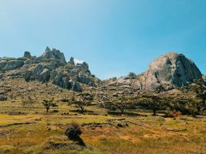 Rocky mountain landscape with large granite formations, scattered trees, grassy plains, and a clear blue sky.